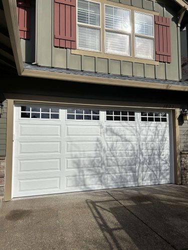 Exterior of a green house with red shutters and stone accents in Portland, showing a white double garage door with windows. Painted by Pearl Painters. Photo by Taylor, April 2025.