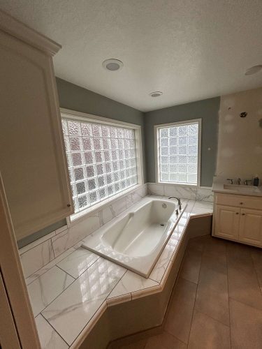This bathroom, painted by Mallen, features a built-in white bathtub surrounded by marble tile, glass block windows, and a sink with cabinets. For your Portland remodel in April 2025, Pearl Painters can help you achieve this look.