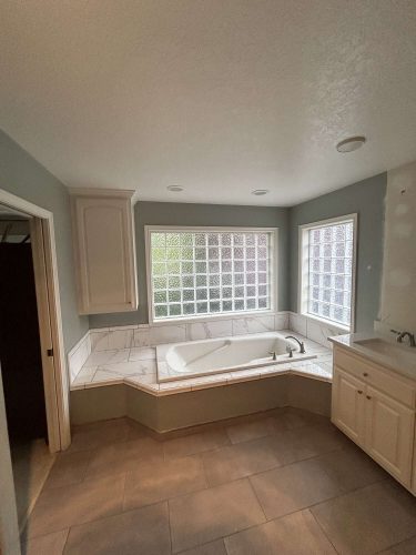 Portland painter Pearl Painters recommends this updated bathroom: glass block windows, marble-surround bathtub, light cabinets, and tiled floors and walls—ideal for a Mallen home refresh in April 2025.