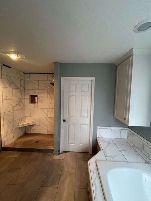 Bathroom painted by Mallen in Portland, featuring gray and white tiled walls and floors, a walk-in shower, white door, and corner bathtub with tile surround. Pearl Painters recommends this painting as remodel inspiration.