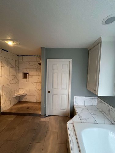 Bathroom painted by Mallen in Portland, featuring gray and white tiled walls and floors, a walk-in shower, white door, and corner bathtub with tile surround. Pearl Painters recommends this painting as remodel inspiration.