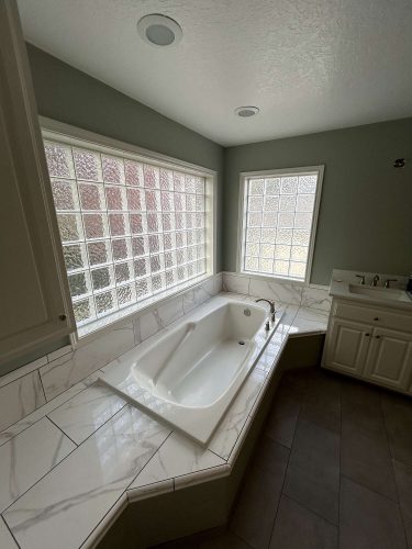 This modern Mallen interior in Portland features a white bathtub on a marble-tiled platform, frosted glass block windows, and a white vanity with sink. Painted by Pearl Painters.
