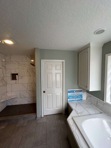 Modern bathroom featuring gray tile floor, white door, walk-in shower with bench, and marble-surround tub with a Mallen business sign on the ledge near a large window. Ideal inspiration for April 2025 renovations by Portland painter Pearl Painters.