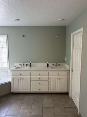 A painter from Pearl Painters completed an updated bathroom in Portland for April 2025, featuring light green painted walls, double-sink white vanity, two soap dispensers, tile floor, and a closed white door.