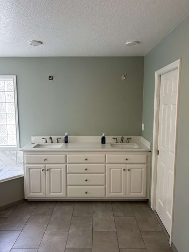 A painter from Pearl Painters completed an updated bathroom in Portland for April 2025, featuring light green painted walls, double-sink white vanity, two soap dispensers, tile floor, and a closed white door.