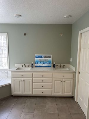 A bathroom vanity with double sinks and white cabinets stands against a light green wall. A Pearl Painters sign is on the countertop. This painting project is in Portland for the Mallen update, April 2025.