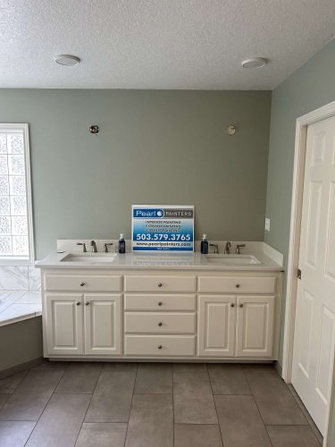 A bathroom vanity with double sinks and white cabinets stands against a light green wall. A Pearl Painters sign is on the countertop. This painting project is in Portland for the Mallen update, April 2025.