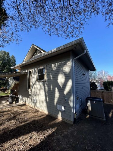 A small light green house in Congdon with a window, exterior air conditioning unit, and shadows from tree branches on the siding under a clear April 2025 sky. Painting by Pearl Painters, Portland painters.