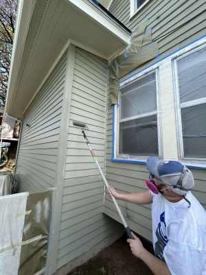 In April 2025, a masked painter from Pearl Painters uses a roller to paint the exterior siding of a Congdon home, with windows taped and covered.