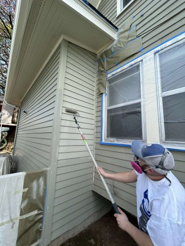 In April 2025, a masked painter from Pearl Painters uses a roller to paint the exterior siding of a Congdon home, with windows taped and covered.