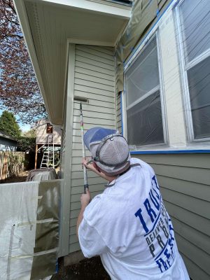 In April 2025, a painter from Pearl Painters refreshes the exterior of a Congdon house in Portland, using a paint roller and covering windows with plastic and tape.