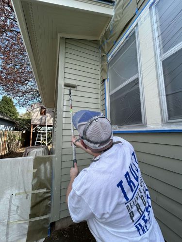 In April 2025, a painter from Pearl Painters refreshes the exterior of a Congdon house in Portland, using a paint roller and covering windows with plastic and tape.