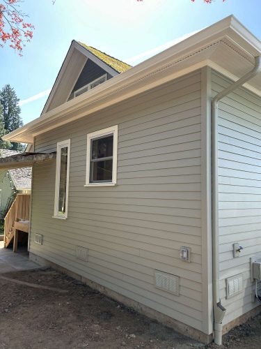 Side view of a light green exterior Congdon house with white trim, two windows, gutter system, and utility boxes. No landscaping around the foundation as of April 2025. Painted by Pearl Painters in Portland.