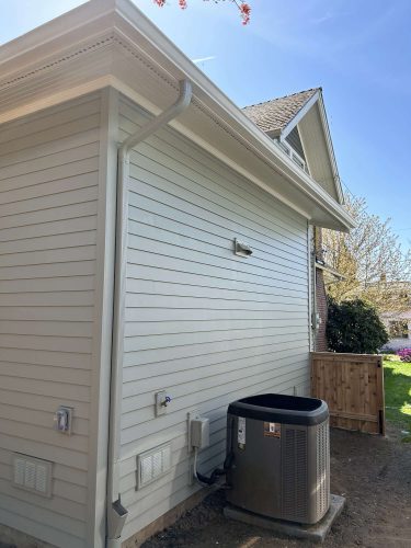 A light-colored vinyl-sided Congdon exterior in Portland with rain gutter, utility boxes, and outdoor HVAC unit on dirt next to a wooden fence, painted by Pearl Painters in April 2025.