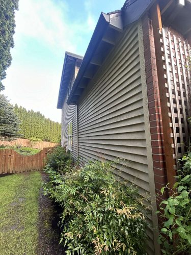 Exterior view of Scott's Portland house in April 2025 after painting by Pearl Painters: beige vinyl siding, brick trim, tall shrubs, wooden fence, and green yard.