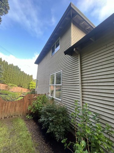 Side view of a beige two-story house in Portland with horizontal siding, two windows, a wooden fence, and green shrubs. Photo by painter Scott for Pearl Painters, April 2025.