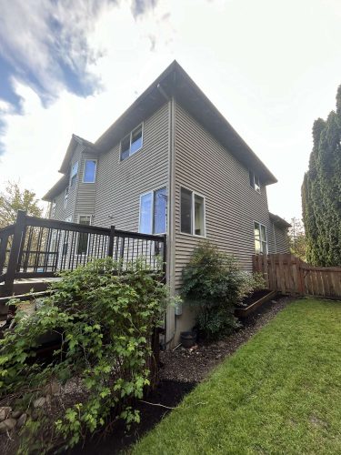 Scott's two-story house in Portland has a beige exterior, multiple windows, a wooden deck, fenced yard, bushes, and a green lawn. Pearl Painters painted the home in April 2025.