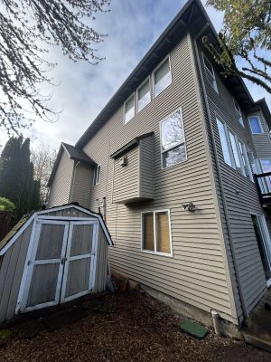 A gray three-story house with horizontal siding and several windows, located next to a weathered wooden shed in a backyard in Portland, is shown. Painting reference photo by painter Scott, April 2025, Pearl Painters.