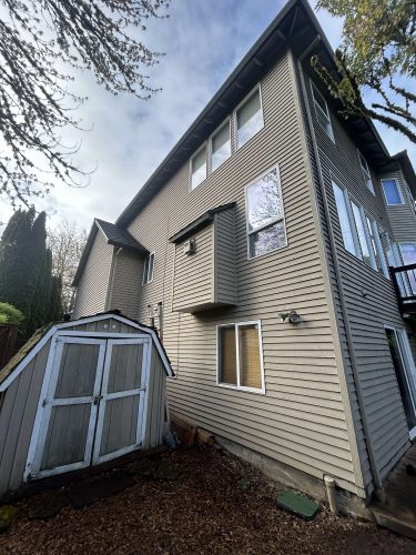 A gray three-story house with horizontal siding and several windows, located next to a weathered wooden shed in a backyard in Portland, is shown. Painting reference photo by painter Scott, April 2025, Pearl Painters.