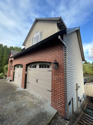 A two-story house in Portland features beige siding and a brick facade, with tan garage doors. Photo taken in April 2025. Painted by Pearl Painters.