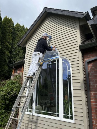 Scott, a painter with Pearl Painters in Portland, stands on a ladder repairing a window on the exterior of a light-colored house with brick trim in April 2025.