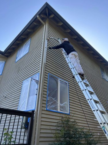 In April 2025, painter Scott paints the exterior trim of a two-story Portland house for Pearl Painters. The windows are covered with protective plastic.