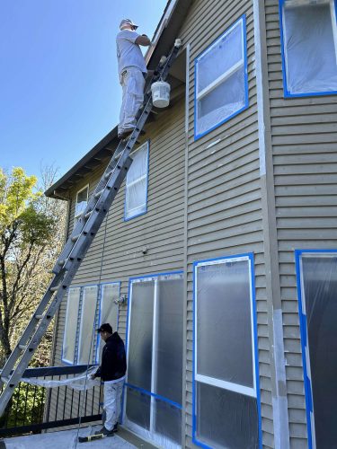In April 2025, Pearl Painters' painters, including Scott, paint the exterior of a house in Portland. One painter works on a ladder near the roof while another stands below. The windows are protected with plastic and blue tape.