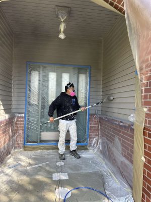 In April 2025, painter Scott from Pearl Painters in Portland spray paints a house exterior, wearing protective gear and using plastic sheets to cover the area.
