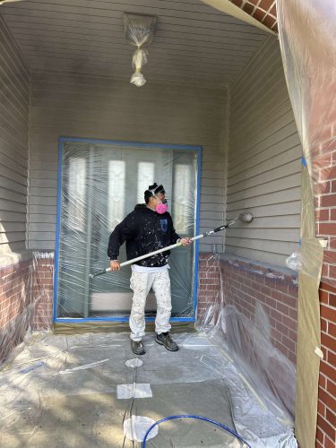 In April 2025, painter Scott from Pearl Painters in Portland spray paints a house exterior, wearing protective gear and using plastic sheets to cover the area.