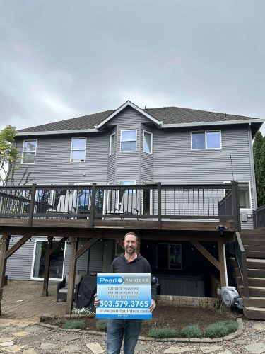 Scott holds a Pearl Painters sign in front of a two-story gray house with a wooden deck in Portland, advertising interior and exterior painting for April 2025.