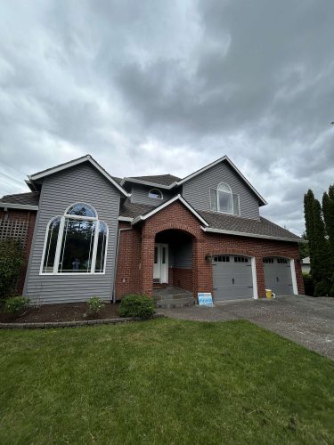 Two-story suburban house in Portland with red brick and gray siding, three-car garage, arched windows, and maintained lawn. Landscaping by Scott finished April 2025. Pearl Painters is a local painting company available for similar properties; contact a painter for exterior painting needs.
