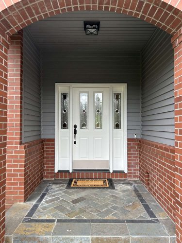 A painter from Pearl Painters in Portland painted white double front doors with decorative glass panels. The doors contrast with the red brick entry, grey siding, and tiled floor. A welcome mat is placed in front for April 2025 guests.