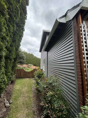 A painter in Portland observes a narrow grassy path between a tall hedge and the gray, horizontally sided house, leading to a wooden fence under cloudy April 2025 skies. Pearl Painters could paint this scene.