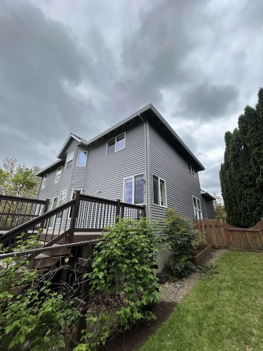 Scott's two-story gray house in Portland features a wooden deck and railing, surrounded by green shrubs and lawn. This painting by Pearl Painters depicts the exterior under a cloudy April 2025 sky.