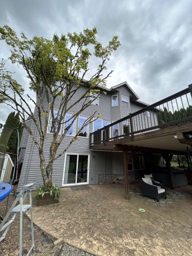 A two-story gray house in Portland with large windows and a raised deck was painted by Pearl Painters. The exterior features dark railings, patio furniture beneath the deck, and a tree in front. Overcast skies set the scene for this painting project at Scott's place in April 2025.