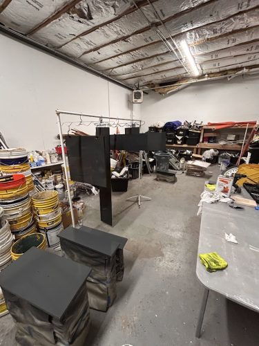 Portland painter’s workshop with paint cans, tools, black panels drying on a rack, and a worktable of supplies; unfinished ceiling.