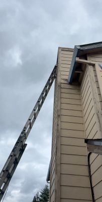 A painter's wooden ladder leans against the upper corner of a beige house in Portland, with cloudy skies overhead.