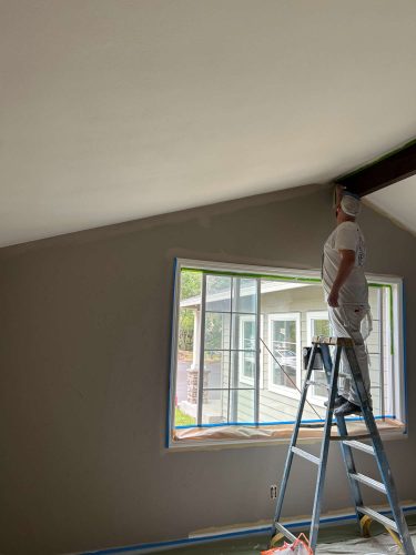 A painter from Pearl Painters stands on a ladder in Portland, painting the upper edge of a living room wall near a window, with painter's tape neatly applied along the trim.