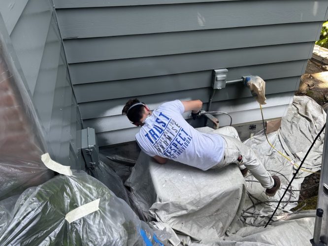 In June 2025, a painter in a white shirt and protective eyewear paints the exterior siding of a house in Carlton, using plastic sheets to protect nearby surfaces. This painting work is not in Portland and is not by Pearl Painters.
