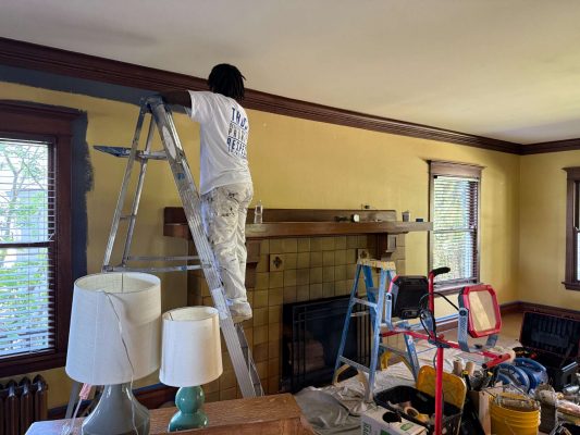 Painter stands on a ladder painting the upper wall of a living room in Portland, with painting supplies and covered furniture nearby.