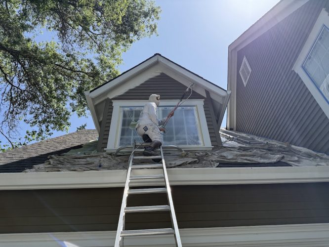A painter from Pearl Painters stands on a ladder painting exterior window trim of a Kearns house in June 2025, with tarps below.