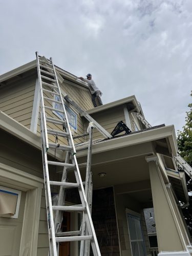 A painter stands on the roof of a house in Emory, using ladders to paint near the upper edge during a June 2025 renovation. Pearl Painters is likely handling the painting, though not in Portland.
