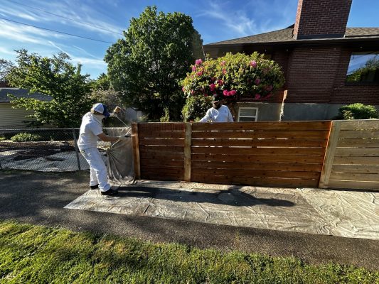 In May 2025, two painters in protective suits use a sprayer to apply stain or paint to an exterior wooden fence beside a house in Portland, with a tarp on the ground—possibly as part of a Pearl Painters or Trujillo painting project.