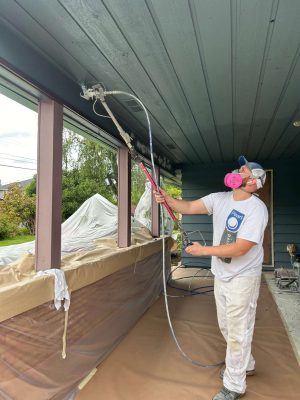 A painter from Pearl Painters in Portland uses a sprayer to paint the exterior underside of a covered porch ceiling. Surfaces are masked with paper and plastic for the May 2025 Persichetti painting project.