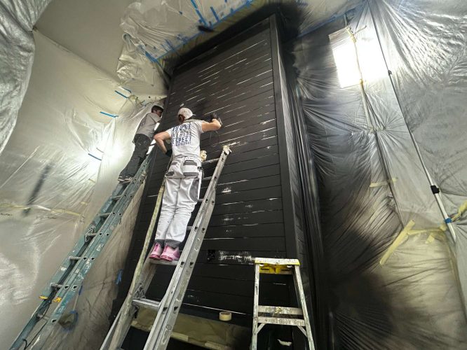 Two painters from the Turk team paint a tall black wooden interior structure in a room covered with plastic sheets. The renovation, managed by Pearl Painters in Portland, is scheduled for completion in May 2025.