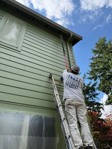 In May 2025, a painter from Pearl Painters in Portland uses a roller to paint the exterior wall of a light green house. The painter wears a white shirt and cap, and plastic covers protect the windows.