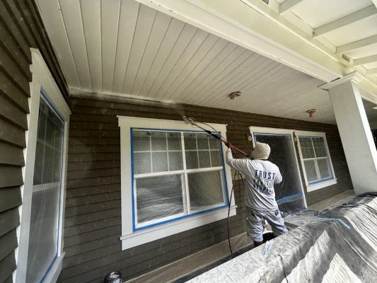 In June 2025, a painter from Pearl Painters uses a sprayer to paint a covered porch ceiling in Kearns; windows and surfaces are taped for protection.
