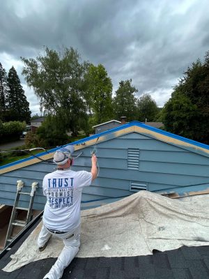 A painter from Pearl Painters is spray painting the upper exterior walls of a blue house for the Persichetti project in Portland, May 2025. The roof is covered with protective sheets and ladders are nearby.