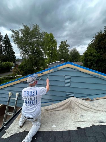 A painter from Pearl Painters is spray painting the upper exterior walls of a blue house for the Persichetti project in Portland, May 2025. The roof is covered with protective sheets and ladders are nearby.