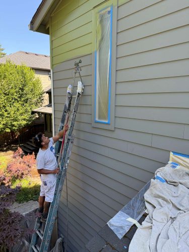 In May 2025, an Arain painter from Pearl Painters paints the exterior siding of a house in Portland, protecting the window with plastic and blue tape.